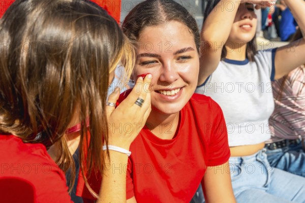 Young women at a sunny stadium, one friend painting red face paint on anothers cheek as they smile and bond, excitedly preparing to cheer for their team