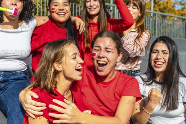Group of excited diverse young women fans cheering loudly and celebrating a sports victory or game while sitting on stadium bleachers with red face paint