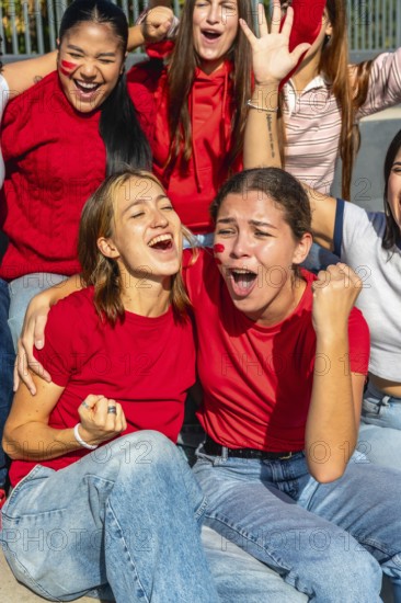 Group of enthusiastic women wearing red shirts and face paint, celebrating with open mouths and raised fists while watching a sporting event from the stadium stands