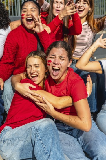 Group of excited young women wearing red, cheering loudly with face paint on their cheeks, embracing and celebrating a victory or a goal with passion and camaraderie
