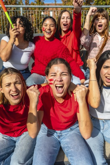 Group of excited young women with face paint and red clothing cheering passionately during a sporting event, celebrating and supporting their team in the stadium
