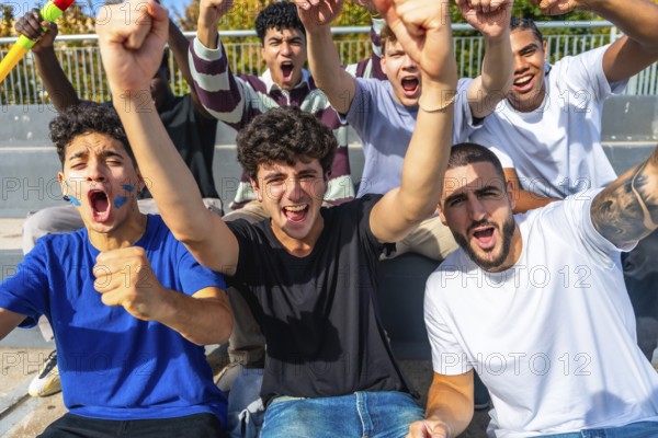 Excited multiethnic male friends cheering and raising fists in outdoor stadium, standing together in bleachers, smiling, shouting and celebrating their teams winning moment