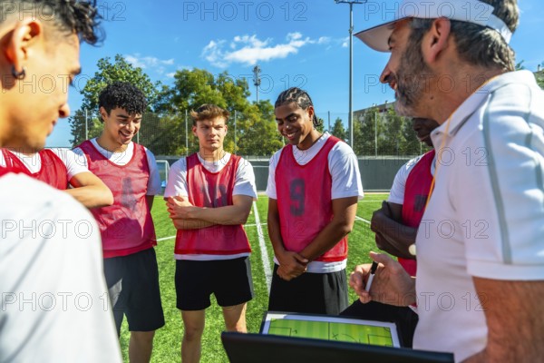 Soccer coach discussing game strategy and explaining tactics on a digital tablet with a group of young male players wearing sports bibs during training on a green field