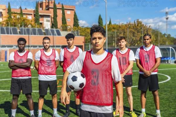 Diverse group of male soccer players in red bibs standing on a training pitch, holding a ball and ready for practice or a competitive match with focused determination