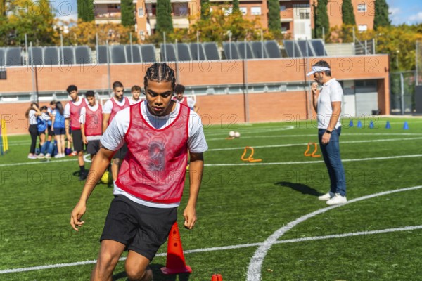 Young male soccer player in a red bib practices drills on green artificial turf with teammates while coach supervises, focusing on technique, movement and teamwork during training