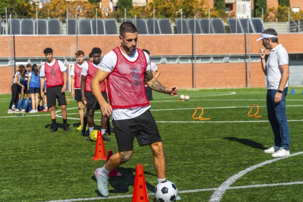 Soccer player in a red bib training on a green artificial turf field, dribbling a football around orange cones while a coach observes the team in the background