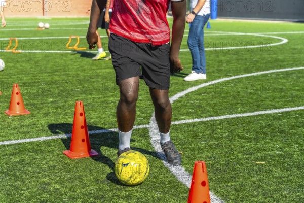 Young player actively training and practicing soccer drills, dribbling the ball around orange cones on a green artificial turf field under natural daylight