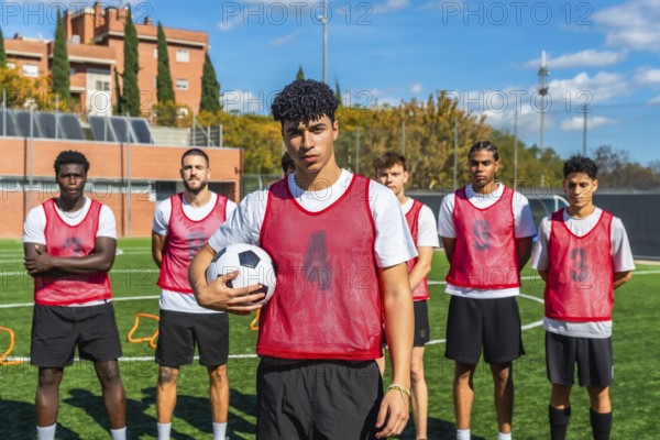 Multi ethnic group of young men in red bibs standing on a green football pitch, preparing for training or a match while showing teamwork and determination