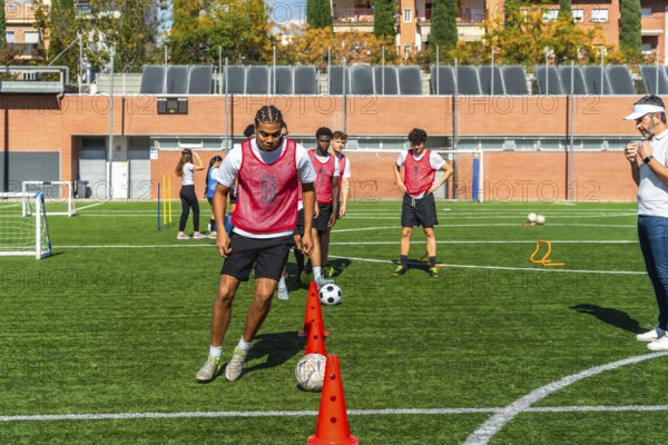 Young men in red vests are performing agility drills with cones and soccer balls on a green artificial turf field, receiving instruction from their coach