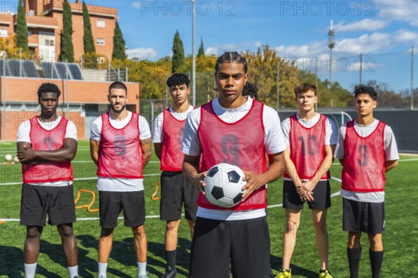 Young men from diverse backgrounds forming a soccer team, standing on a green artificial turf field with a player holding the ball, training for their sport