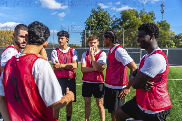 Young male soccer players wearing red bibs huddling on a green artificial turf field, listening intently to their coach or teammate, actively planning their game strategy and building team spirit