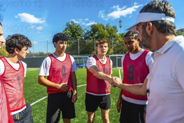 Coach wearing a visor and whistle shaking hands with a young male soccer player on a green football field, with teammates watching during a training session