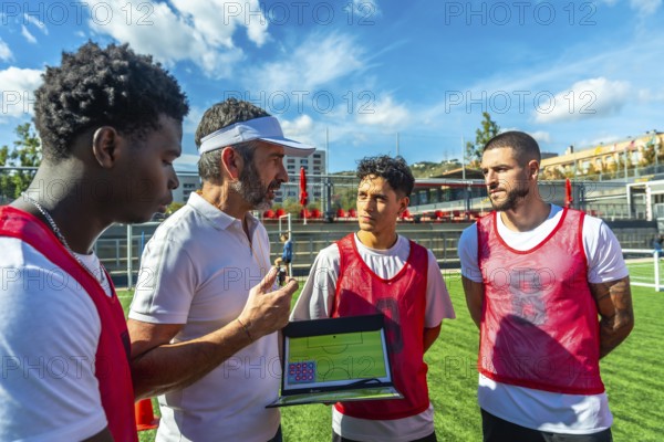 Soccer coach discussing game strategy with diverse football players on a sunny training field, using a tactical board to illustrate positioning and team plays