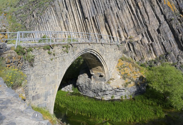 Nature surrounds an old stone bridge, which crosses a densely vegetated river and is flanked by rocks, Melik Tangi bridge, behind it a basalt column formation, Vorotan, Syunik province, Armenia