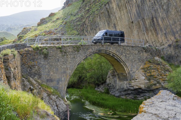 A black van drives over an old stone bridge, surrounded by picturesque rocky and river landscape, camper, motorhome on the Melik Tangi Bridge, behind a basalt column formation, Vorotan River, Syunik Province, Armenia