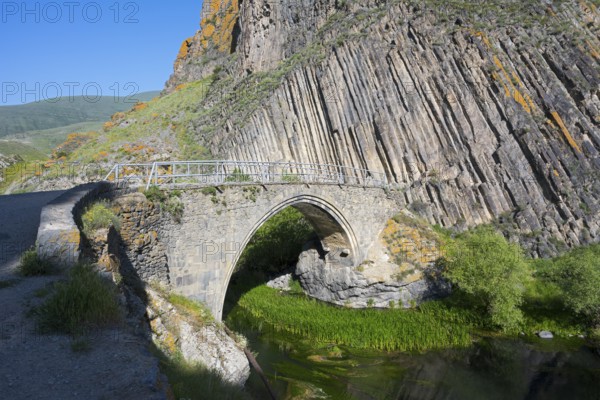 An old stone bridge stands in front of dramatic rock walls surrounded by green vegetation and a blue sky, Melik Tangi Bridge, behind it a basalt column formation, Vorotan River, Syunik Province, Armenia