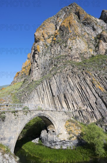 A high stone bridge stretches in front of an impressive rock formation under a bright blue sky, Melik Tangi Bridge, behind it a basalt column formation, Vorotan River, Syunik Province, Armenia
