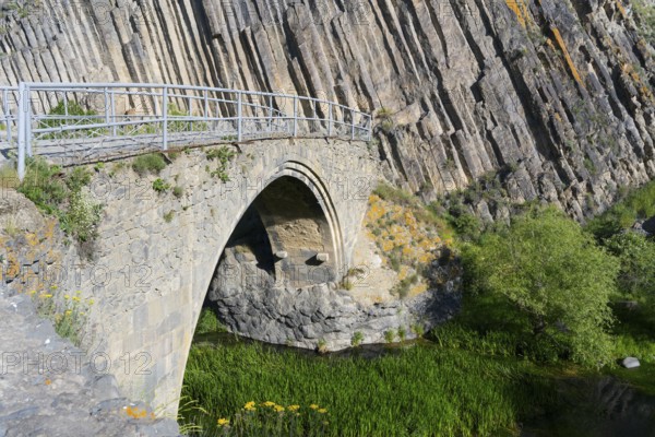 View of a stone bridge across a clear river with lush greenery and dramatic rock formations in the background, Melik Tangi Bridge, behind a basalt column formation, Vorotan River, Syunik Province, Armenia