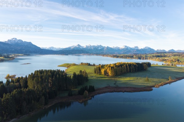 Wide alpine landscape with lake and wooded hills in the morning light, surrounded by snow-capped mountains