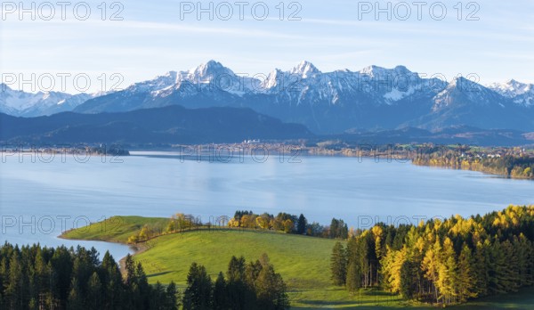 Alpine panorama with snow-capped peaks, calm lake and autumn-colored trees under a blue sky
