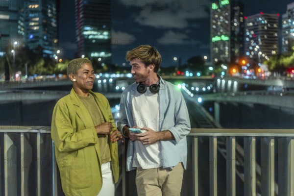 Diverse young couple chatting and smiling on an urban bridge at night, city skyline and glowing streetlights framing a candid, modern moment of connection and joy