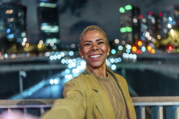 Happy black woman capturing a selfie at an urban bridge, her bright smile and short hair illuminated by the vibrant bokeh lights of the bustling city nightlife