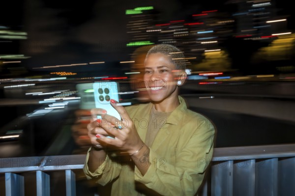 Woman smiling and holding a smartphone, capturing the vibrant motion blur of city lights at night, creating a dynamic urban scene with long exposure light trails