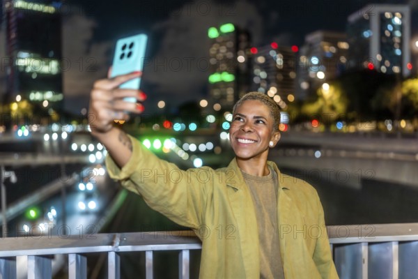 Woman with short blonde hair and happy expression capturing a selfie with her smartphone against a vibrant urban cityscape at night, showcasing technology and connection after dark