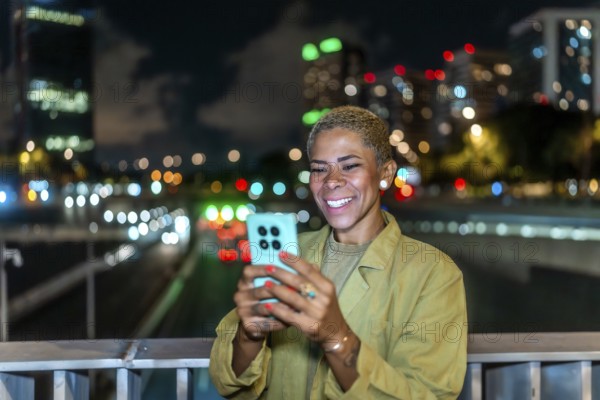 Woman smiling while interacting with her mobile phone, standing on a bridge with blurred city lights and traffic creating a bokeh effect in the urban background at night