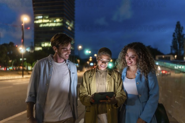 Diverse group of young adults walking a city sidewalk at night, smiling and sharing content on a glowing tablet as street lights and architecture blur into a warm urban bokeh