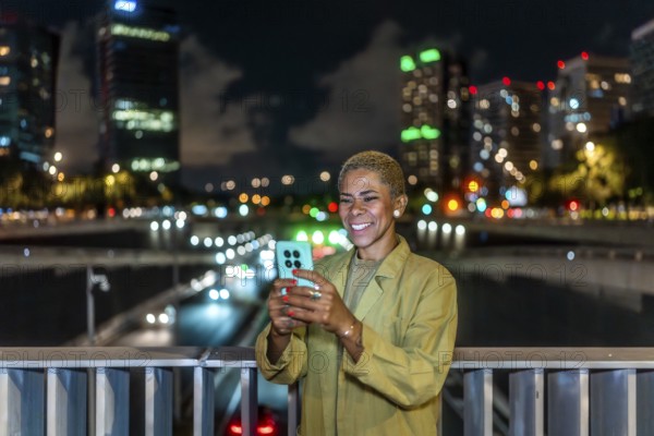 Woman standing on a city bridge at night, smiling while using a smartphone with blurred traffic and illuminated buildings creating a warm bokeh background