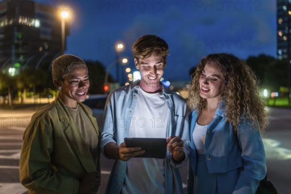 Three diverse friends, two women and one man, standing on a city street at night, smiling and enjoying social connection while looking at content on a shining tablet