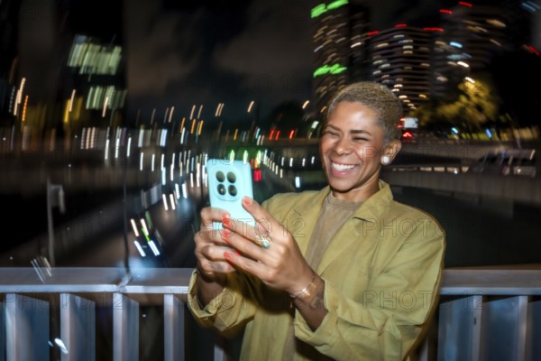 Smiling woman with short blond hair takes a nighttime smartphone selfie in an urban setting, bokeh city lights and blurred buildings creating a joyful, modern travel moment