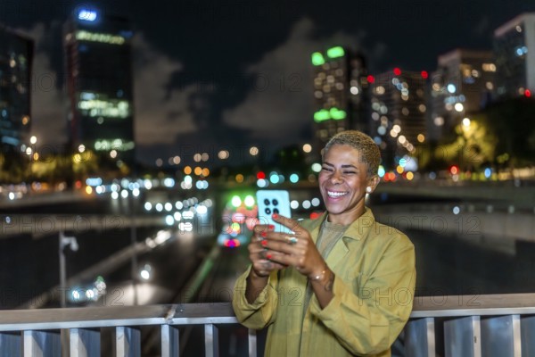 Happy woman smiling, holding a smartphone and enjoying a vibrant urban night view from a bridge, with illuminated city buildings and bokeh lights in the background