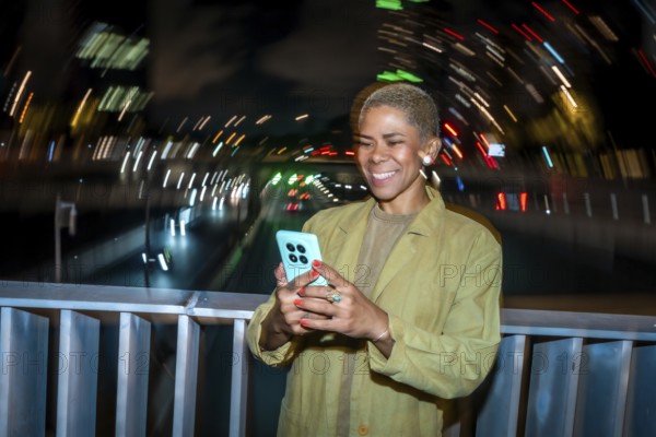 Smiling young adult woman using a mobile phone at night on a city bridge, enjoying digital connectivity with blurred light trails and urban movement in the background