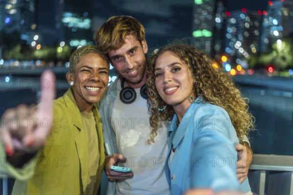 Three happy diverse friends are taking a selfie together, smiling at the camera and celebrating friendship against a vibrant blurred city lights background at night