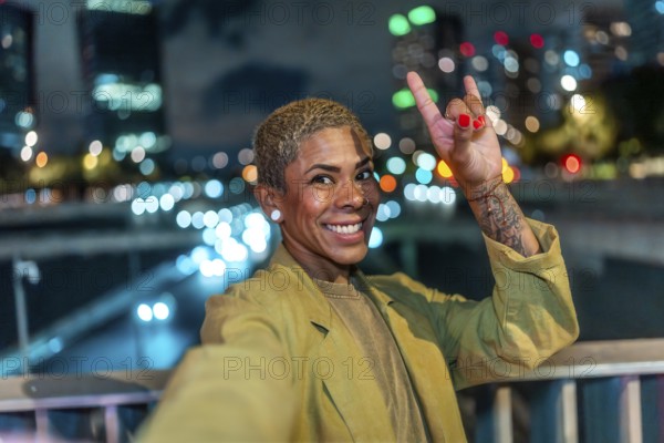 Happy woman with short blond hair and a tattoo making a rock and roll hand gesture, smiling at the camera during a vibrant night out with city lights bokeh in the background