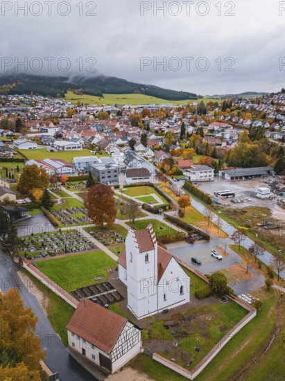 View of a town with church and cemetery surrounded by autumn trees and residential buildings on a cloudy day, Wehingen, Tuttlingen district, Germany