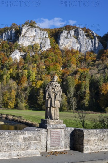 Statue, saint statue of Saint Nepomuk, patron saint of bridges, bridge saint on the historic arched bridge, Hausener Brücke behind the distinctive Hausener Zinnen, climbing rocks, Jurassic limestone rocks, Weissjura, Jurassic rocks, limestone rocks, surrounded by autumnal vegetation, Hausen im Tal, Upper Danube Valley, Baden-Württemberg, Germany