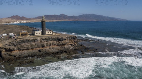 Aerial view of Faro Punta de Jandia lighthouse at the southern tip of Jandia peninsula, in the foreground white foam crowns on moving sea swell large high waves on reef edge beaming sea strong surf, Fuerteventura, Canary Islands, Canary Islands, Spain