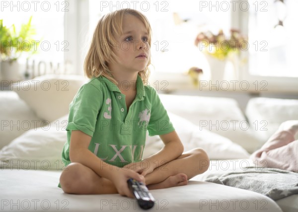 Child, boy, 7 years old, blonde, sitting on sofa, with remote control in hand, watching TV, watching television, watching television, excited, fascinated, Stuttgart, Baden-Württemberg, Germany