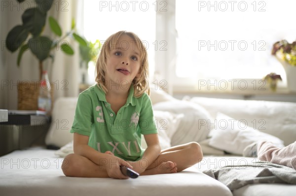 Child, boy, 7 years old, blonde, sitting on sofa, with remote control in hand, watching TV, watching television, in front of television, playing with tongue, Stuttgart, Baden-Württemberg, Germany