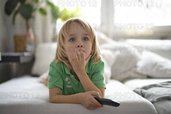 Child, boy, 7 years old, blonde, lying on sofa with remote control in hand, watching TV, watching television in front of television, excited, fascinated, Stuttgart, Baden-Württemberg, Germany