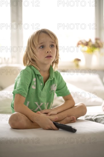 Child, boy, 7 years old, blonde, sitting on sofa, with remote control in hand, watching TV, watching television, watching television, excited, fascinated, Stuttgart, Baden-Württemberg, Germany