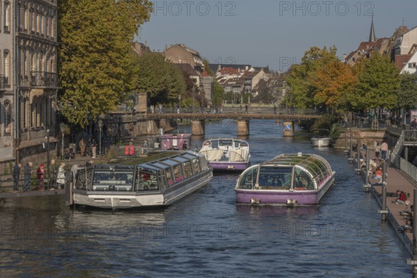 Tourist boat docks on the Ill and transports visitors who enjoy the view of Strasbourg's sights and trees in autumn. The scenery is peaceful and sunny. Bas Rhin, Alsace, France