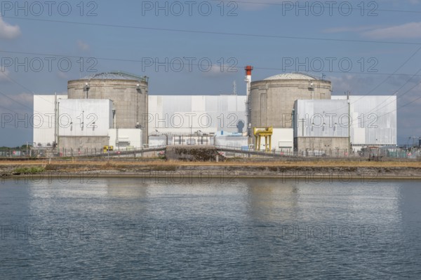 A large industrial nuclear power plant is located on a quiet water front. Two tall cylindrical structures dominate the skyline, with overhanging power lines and distant clouds that ennoble the blue sky. Fessenheim, Haut Rhin, Alsace, France