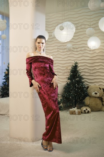 A woman wearing a luxurious red off-shoulder gown stands confidently in a tastefully decorated indoor space