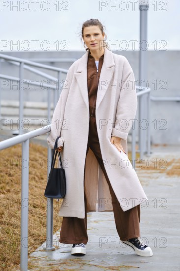 A woman stands confidently on outdoor steps, wearing a long light coat over a brown outfit. She carries a black bag and wears sneakers, with a gray sky visible above her