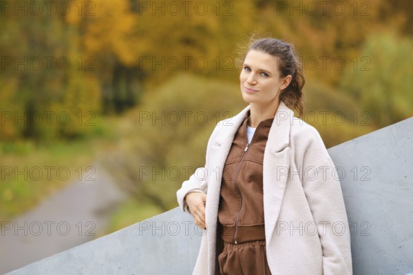 A woman with long hair stands confidently against a gray geometric structure. She wears a brown outfit and smiles softly. The background features warm autumn foliage and a pathway
