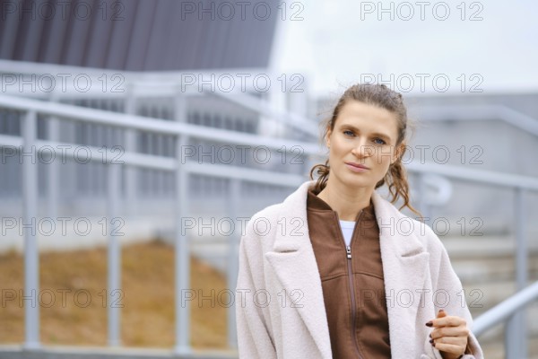 A woman stands confidently outside near contemporary buildings. She wears a fashionable coat and has a natural expression. The sky is overcast, creating a calm atmosphere in the background
